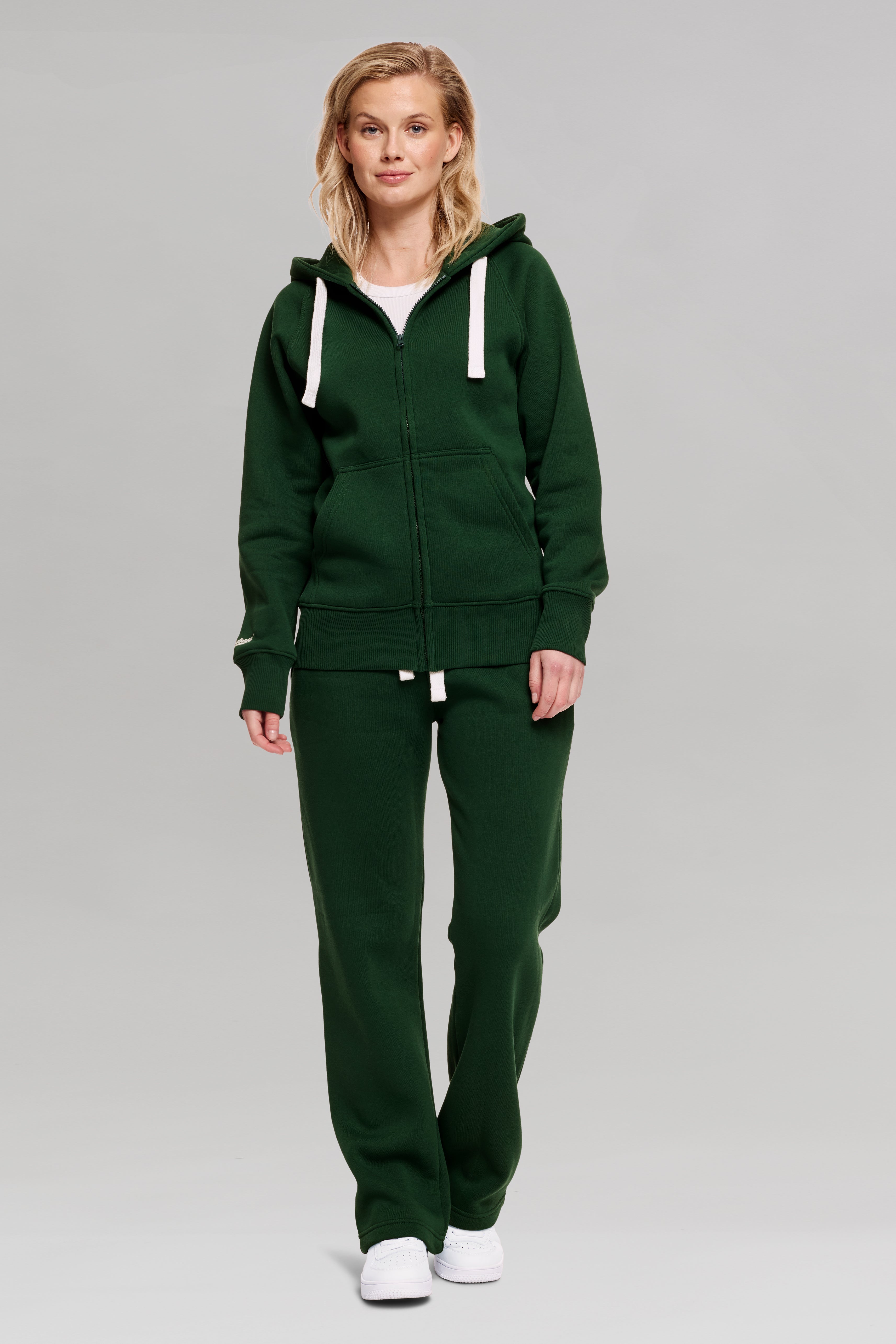 A female model wearing a green hoodzip jacket and matching green sweatpants with a large kangaroo pocket, white hoodie strings, and rib at bottom hem and cuffs.